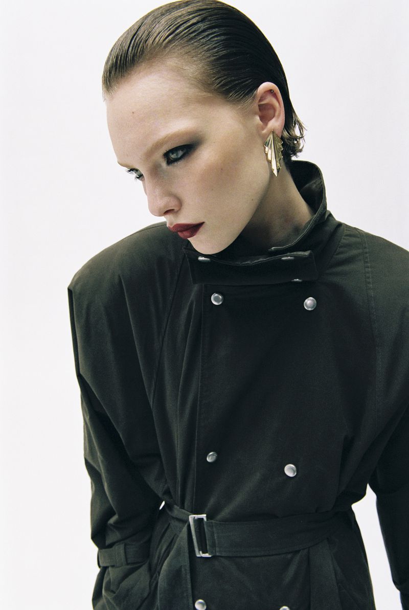 A striking portrait of a woman with slicked-back hair, dark eye makeup, and a dark belted coat against a clean white backdrop.