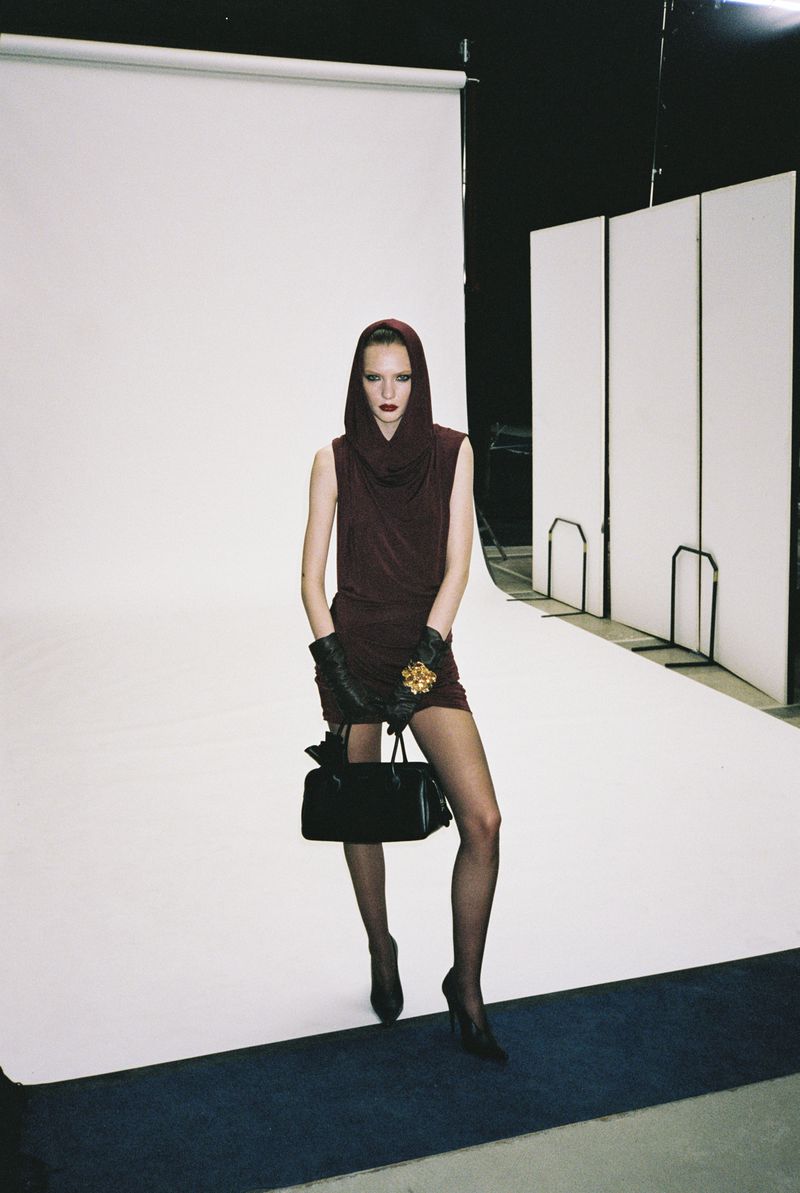 A female fashion model stands in a photo studio wearing a dark burgundy dress, matching head covering, gloves, and carrying a purse.