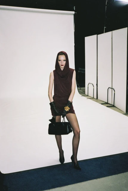 A female fashion model stands in a photo studio wearing a dark burgundy dress, matching head covering, gloves, and carrying a purse.