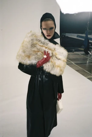 Model wearing a black leather coat with a voluminous fur stole, red leather gloves, and a black headpiece in a photo studio.