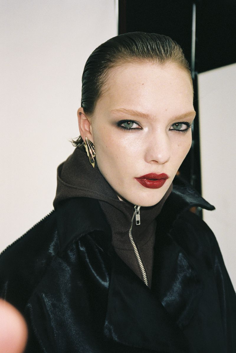 Portrait of a fashion model in a dark hooded jacket with red lipstick, smokey eyeshadow, and gold earrings against a neutral background.