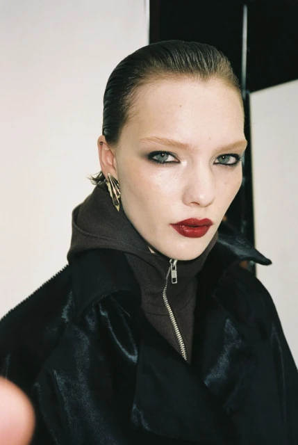 Portrait of a fashion model in a dark hooded jacket with red lipstick, smokey eyeshadow, and gold earrings against a neutral background.