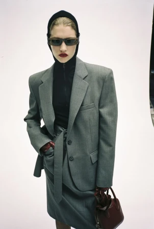 Model wearing an oversized grey tailored suit with a black hood, dark sunglasses, and burgundy accessories against a white background.