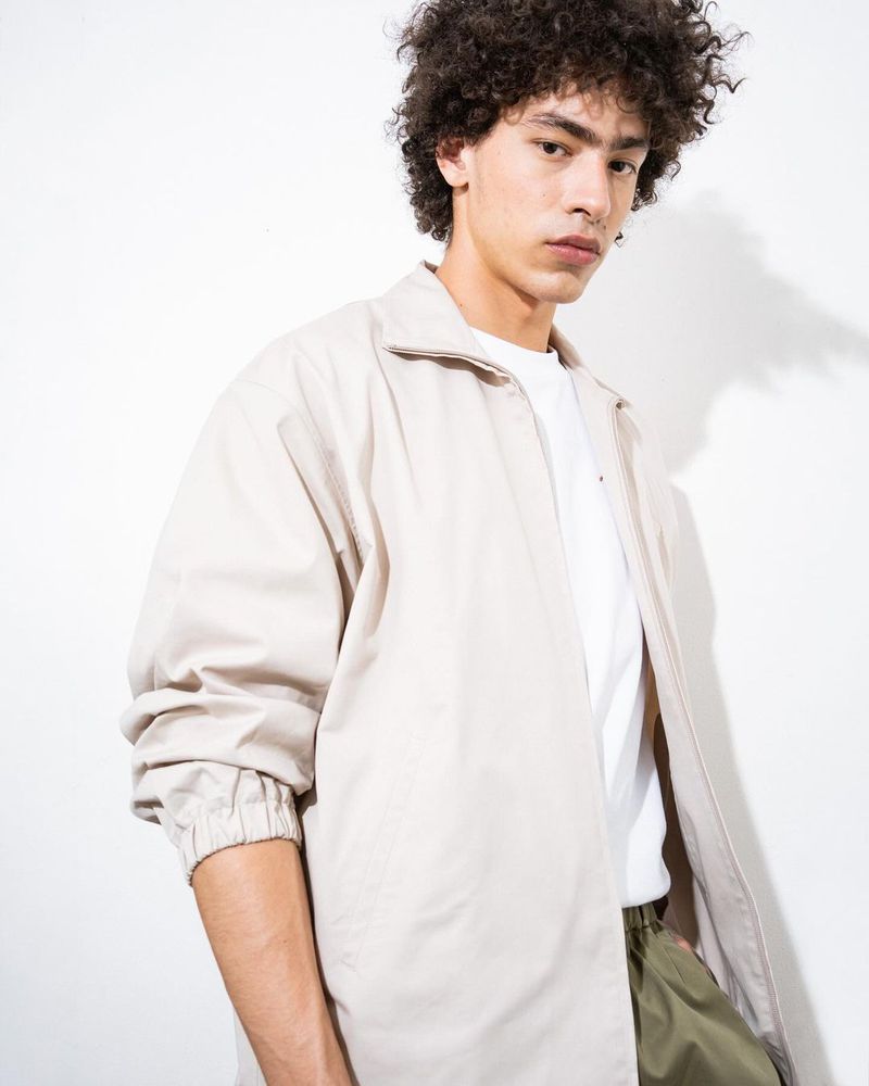 Young man with curly hair poses in casual jacket against a white background, showcasing minimalist fashion and neutral color palette.