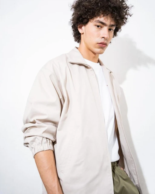 Young man with curly hair poses in casual jacket against a white background, showcasing minimalist fashion and neutral color palette.