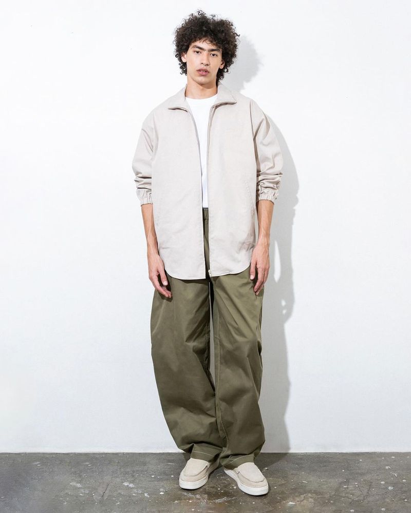 Full body shot of a young adult male wearing a white t-shirt, beige zip-up jacket, oversized olive pants, and loafers standing in a studio.
