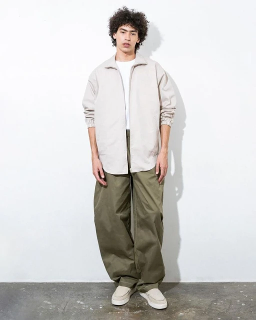 Full body shot of a young adult male wearing a white t-shirt, beige zip-up jacket, oversized olive pants, and loafers standing in a studio.