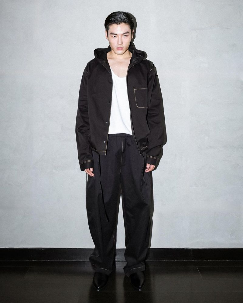 A fashionable male model stands in a photo studio wearing a black hooded jacket, white tank top, and black pants against a grey wall.