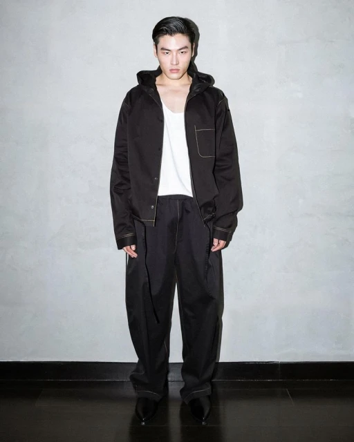 A fashionable male model stands in a photo studio wearing a black hooded jacket, white tank top, and black pants against a grey wall.