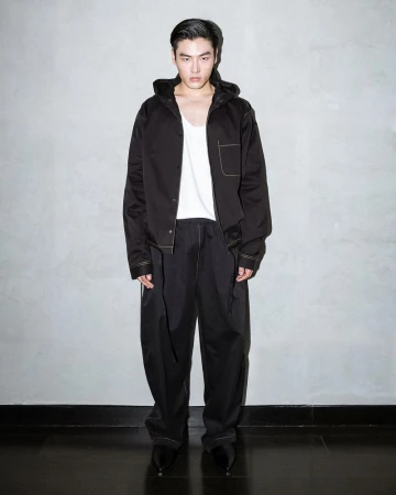 A fashionable male model stands in a photo studio wearing a black hooded jacket, white tank top, and black pants against a grey wall.