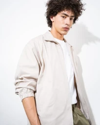 Male model with curly hair wearing an open beige zip jacket over a white shirt against a white backdrop.