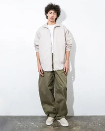 Model wearing a light beige zip jacket and baggy olive green trousers against a white studio wall.