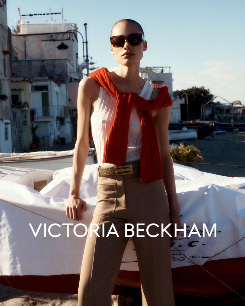 Fashionable adult woman poses near a boat wearing sunglasses, a ribbed tank top, tan pants, and a red sweater tied around her neck.