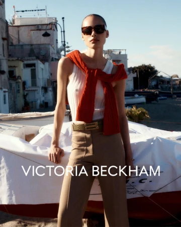Fashionable adult woman poses near a boat wearing sunglasses, a ribbed tank top, tan pants, and a red sweater tied around her neck.