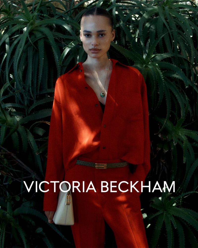 Fashion shot of a young woman wearing a red shirt and pants with a cream-colored handbag, posing in front of lush green plants.