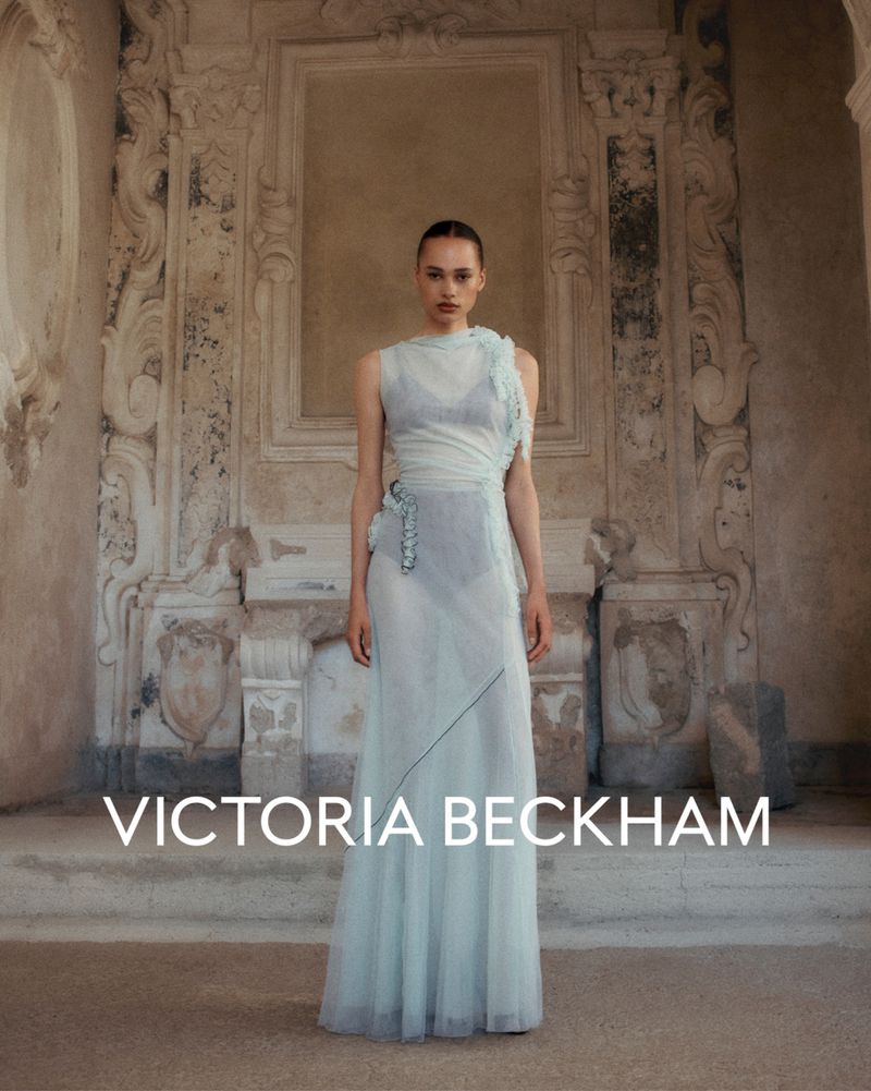A model poses in a light blue, sheer dress in front of an ornate, aged wall with architectural details for a fashion editorial.