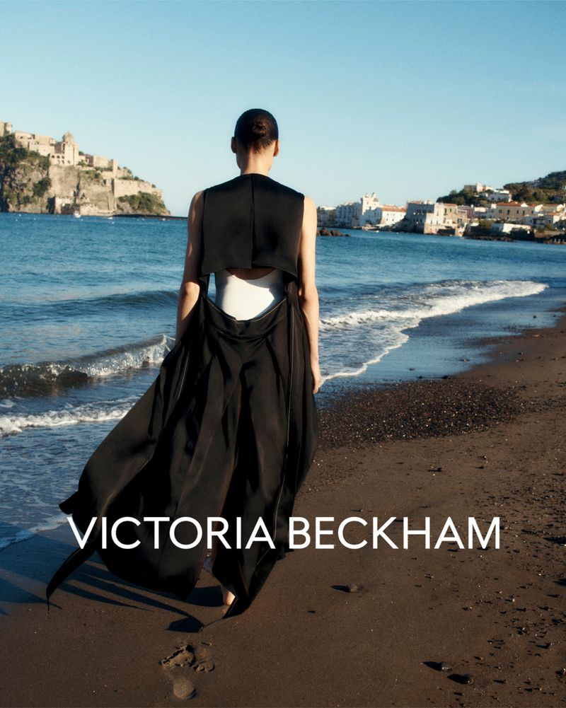 Model Annemary Aderibigbe seen from behind walking along a dark sand beach in a black open-back dress for Victoria Beckham.