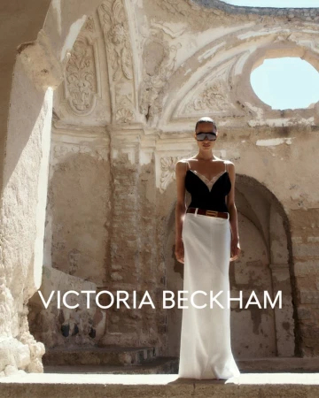 Victoria Beckham stands outdoors against an old building, wearing a black top, white skirt, brown belt, and sunglasses in an eye-level shot.