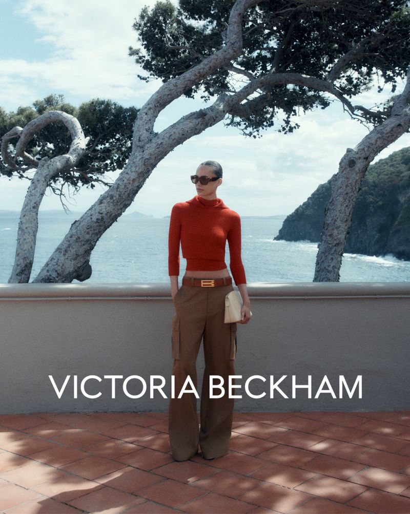 A woman with sunglasses stands on a brick terrace with a seascape background. She is wearing a brown top and wide-leg trousers.