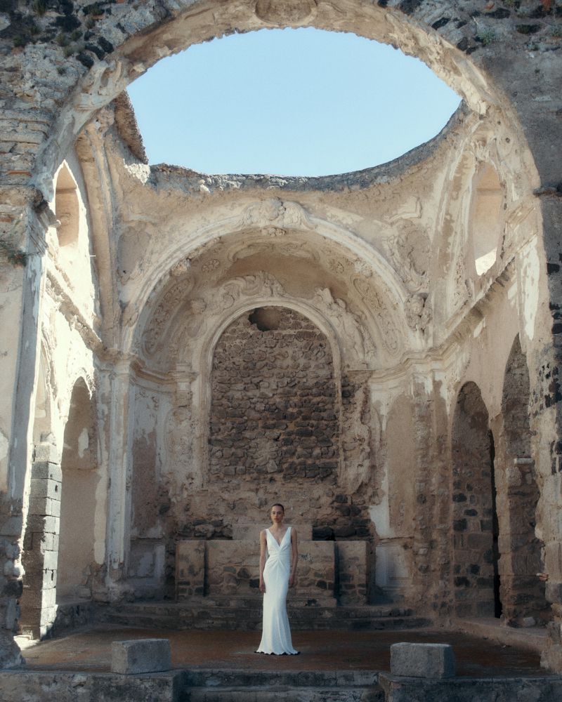 An adult female in a long white dress stands inside the decaying stone ruins of an old building on a sunny day with a clear blue sky.