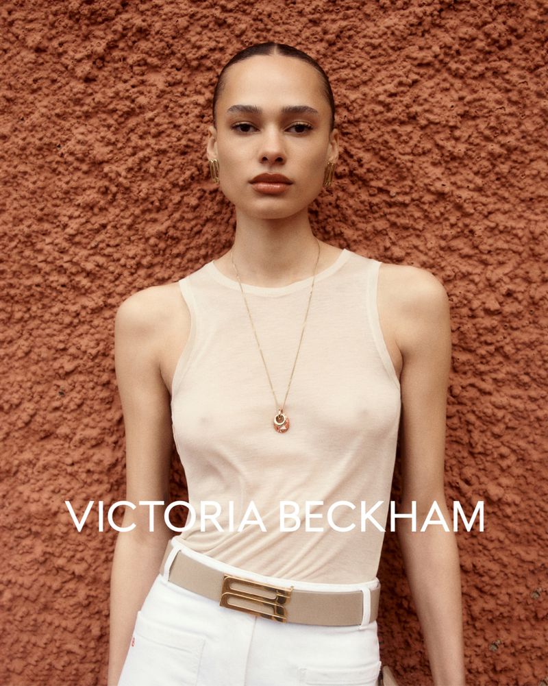 Portrait of a fair-skinned woman with slicked-back hair, wearing a sheer top, white pants, and a gold necklace, posing against an textured orange wall.