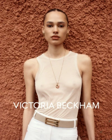 Portrait of a fair-skinned woman with slicked-back hair, wearing a sheer top, white pants, and a gold necklace, posing against an textured orange wall.