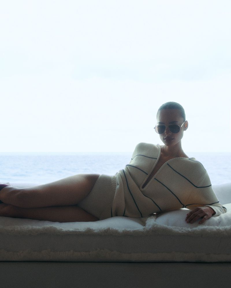 Woman in sunglasses poses in a knitted sweater and briefs, lying on a white surface with an ocean background.