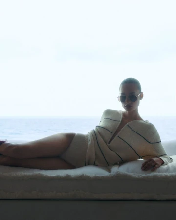 Model Annemary Aderibigbe reclines on a white daybed wearing sunglasses and a cream striped knit sweater overlooking the ocean.