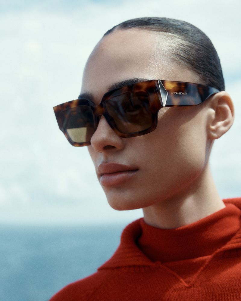 A woman in a red sweater wears large, square, tortoiseshell sunglasses against a blue sky and water backdrop in a fashion editorial.