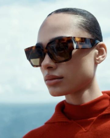 A woman in a red sweater wears large, square, tortoiseshell sunglasses against a blue sky and water backdrop in a fashion editorial.