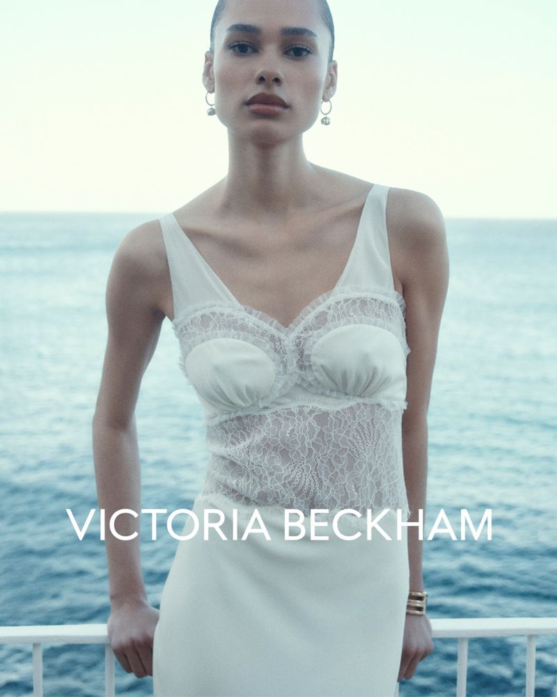 Woman in a cream-colored dress with lace bodice and silver earrings stands near a white railing overlooking the ocean on a clear day.