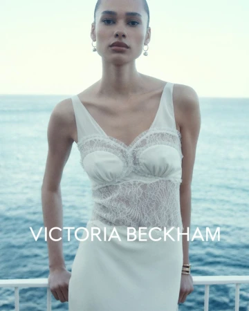 Woman in a cream-colored dress with lace bodice and silver earrings stands near a white railing overlooking the ocean on a clear day.