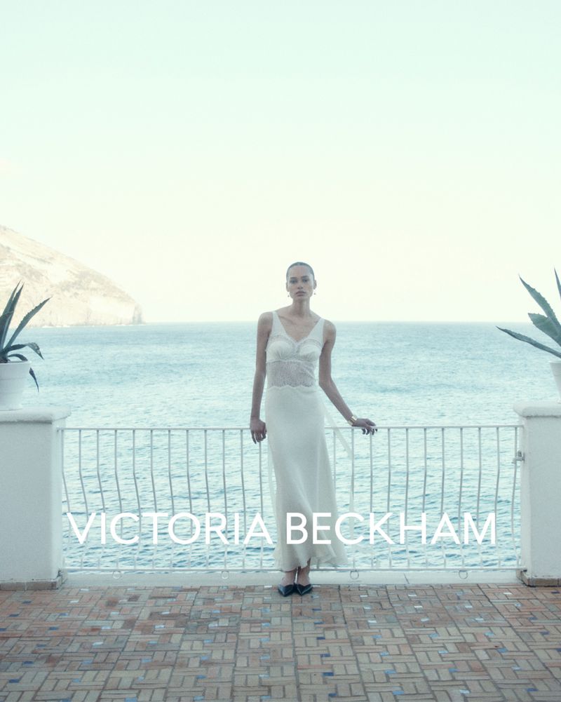 A woman in a white dress stands on a tiled terrace overlooking the ocean on a bright day, with plants and a railing visible.