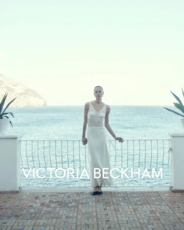 A woman in a white dress stands on a tiled terrace overlooking the ocean on a bright day, with plants and a railing visible.