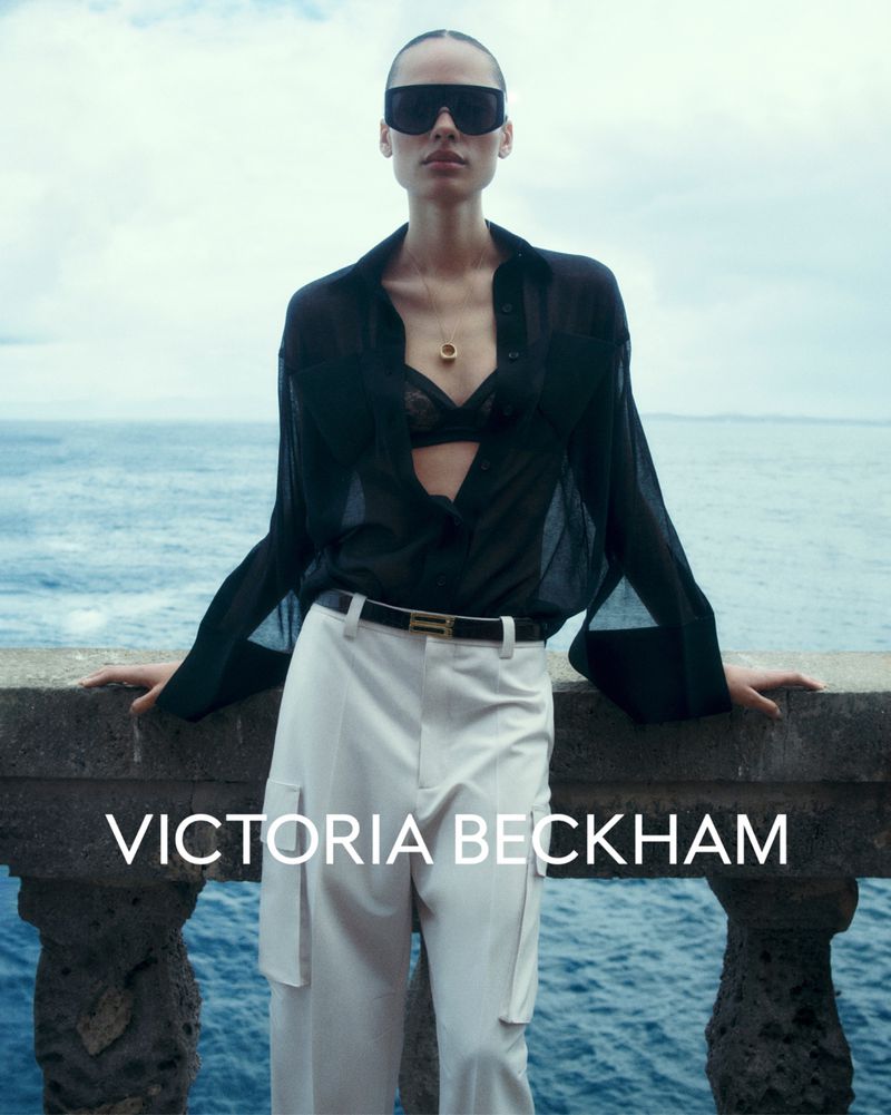Fashion shot of an adult female wearing sunglasses, a black unbuttoned blouse, and white pants. The ocean is visible in the background.