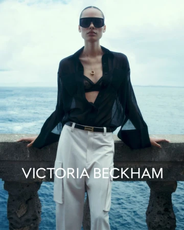 Fashion shot of an adult female wearing sunglasses, a black unbuttoned blouse, and white pants. The ocean is visible in the background.
