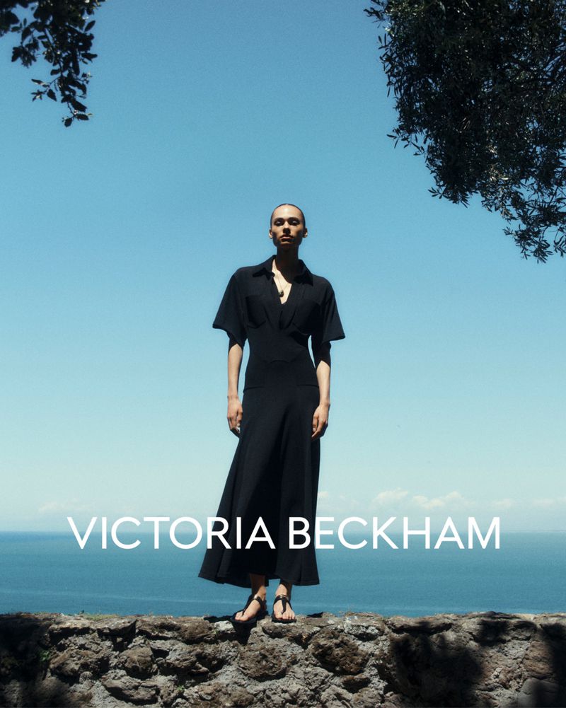A person stands on a stone structure overlooking the ocean, modeling a black dress by Victoria Beckham under a clear blue sky.