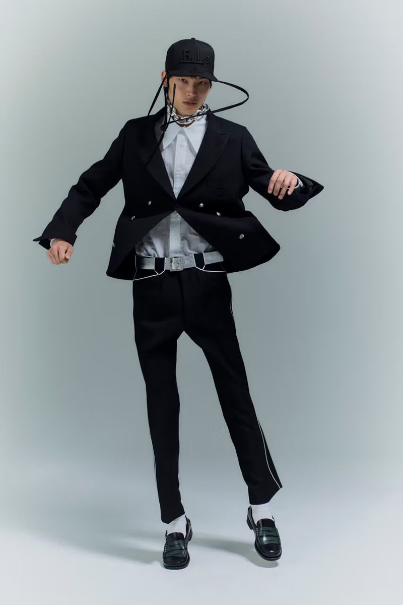 Fashionable adult male wearing a black blazer, pants, and baseball cap poses dynamically in a studio with soft, diffused lighting.