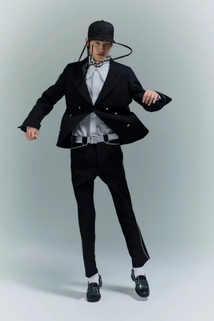 Fashionable adult male wearing a black blazer, pants, and baseball cap poses dynamically in a studio with soft, diffused lighting.