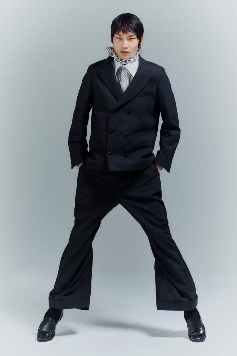 Young male model in a black double-breasted suit with wide-leg trousers, posing against a plain grey studio backdrop, fashion editorial.
