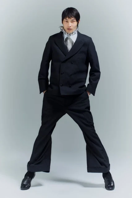 Young male model in a black double-breasted suit with wide-leg trousers, posing against a plain grey studio backdrop, fashion editorial.