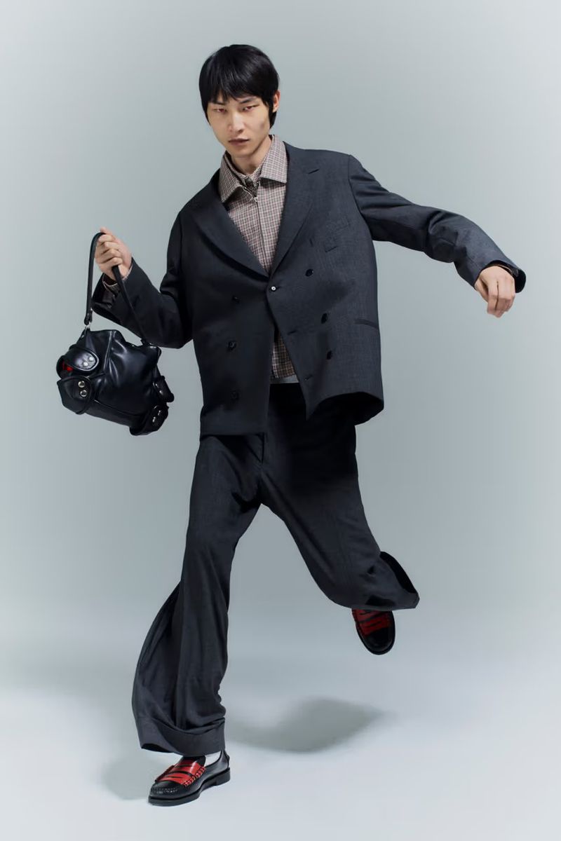 Model wearing an oversized dark grey suit and holding a black leather bag in dynamic motion against a light studio backdrop.