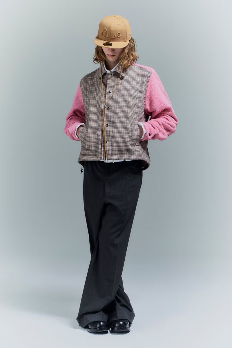 A person in a beige baseball cap, pink and tan plaid jacket, and black pants poses in a full shot against a pale gray background.