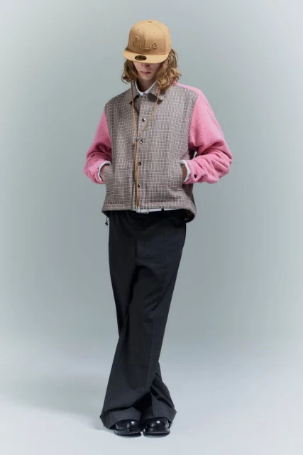A person in a beige baseball cap, pink and tan plaid jacket, and black pants poses in a full shot against a pale gray background.