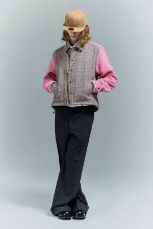 A person in a beige baseball cap, pink and tan plaid jacket, and black pants poses in a full shot against a pale gray background.