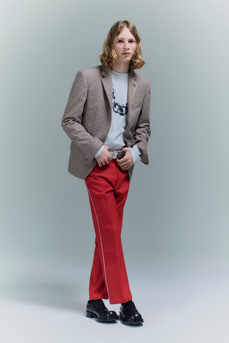 Full-length shot of a young adult male posing indoors wearing a blazer, sweater, red trousers with a stripe, and black shoes.