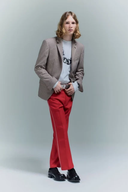 Full-length shot of a young adult male posing indoors wearing a blazer, sweater, red trousers with a stripe, and black shoes.