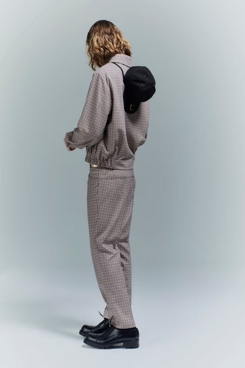 Model seen from behind wearing a patterned grey jacket and matching trousers with a black cap on the back in a studio setting.