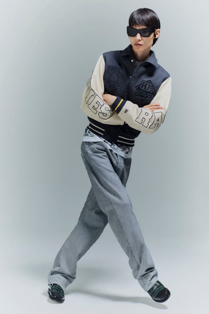 Model in dark sunglasses wearing a two-tone varsity jacket and wide-leg grey trousers against a light grey studio background.