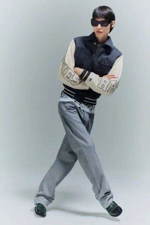 Model in dark sunglasses wearing a two-tone varsity jacket and wide-leg grey trousers against a light grey studio background.